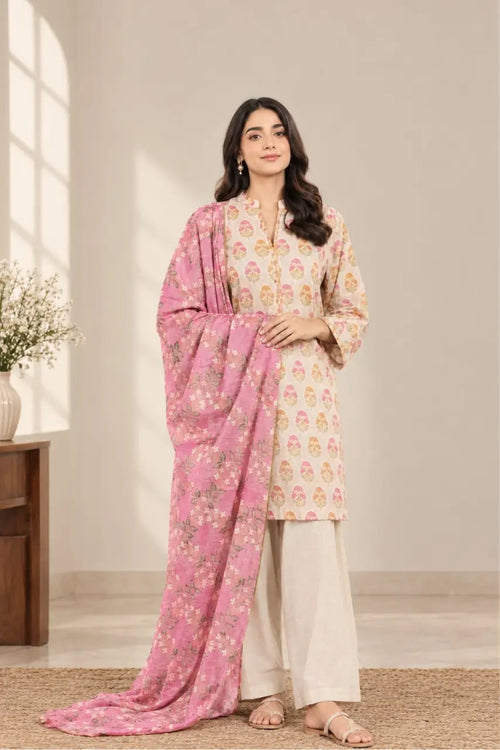A woman models a traditional salwar kameez suit set featuring a cream-colored kurta with pink and gold floral prints, cream-colored palazzo pants, and a pink dupatta with gold floral motifs, set against a warm beige background.