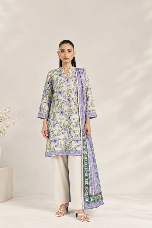 A woman models a three-piece traditional outfit featuring a light green kurta with purple and green floral patterns, paired with light beige wide-leg trousers and a matching purple patterned dupatta, set against a neutral beige background.