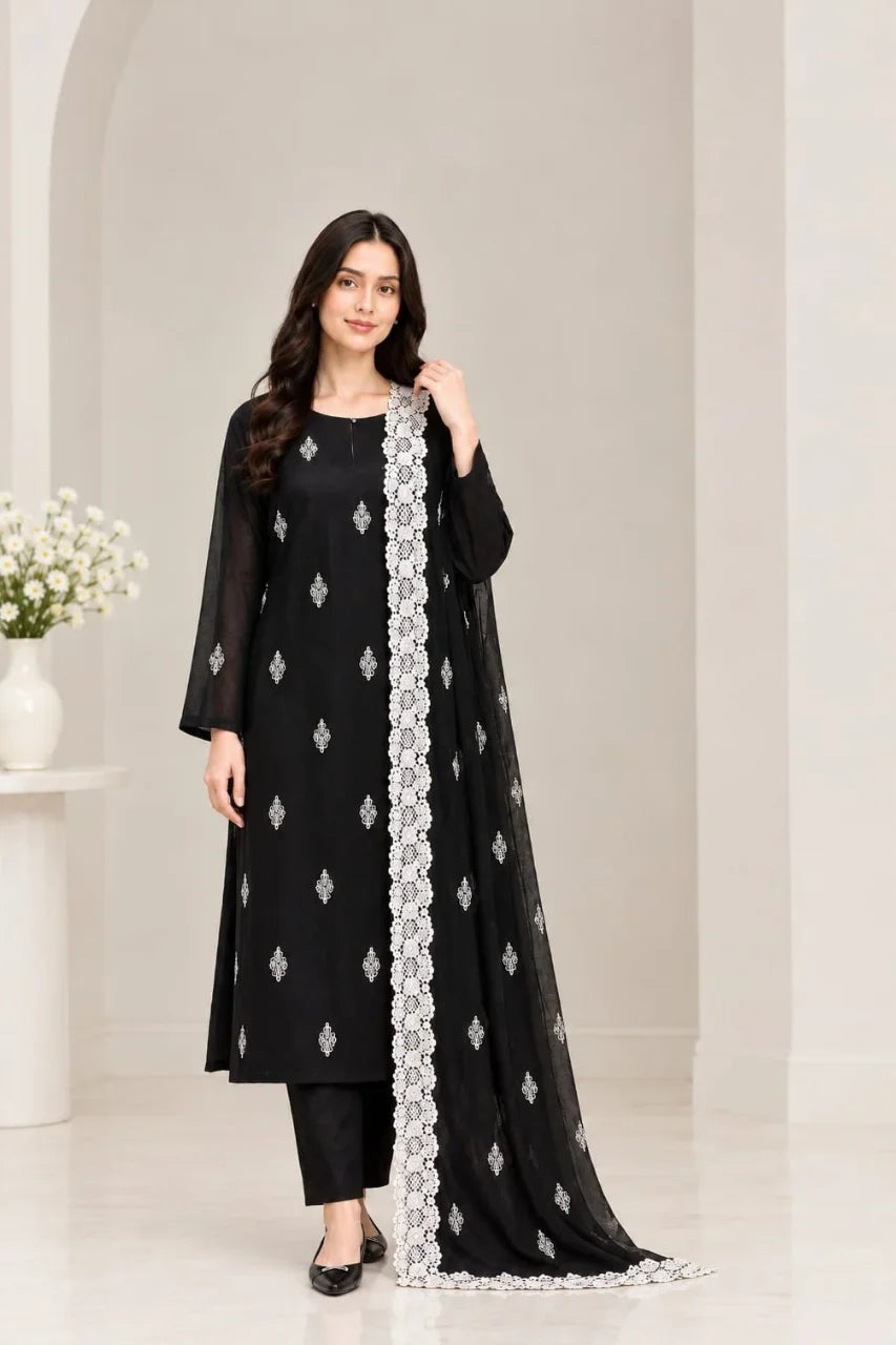 A woman modeling a black three-piece traditional outfit with silver embroidered patterns and a matching dupatta draped over one shoulder, set against a minimalist cream-colored interior background.