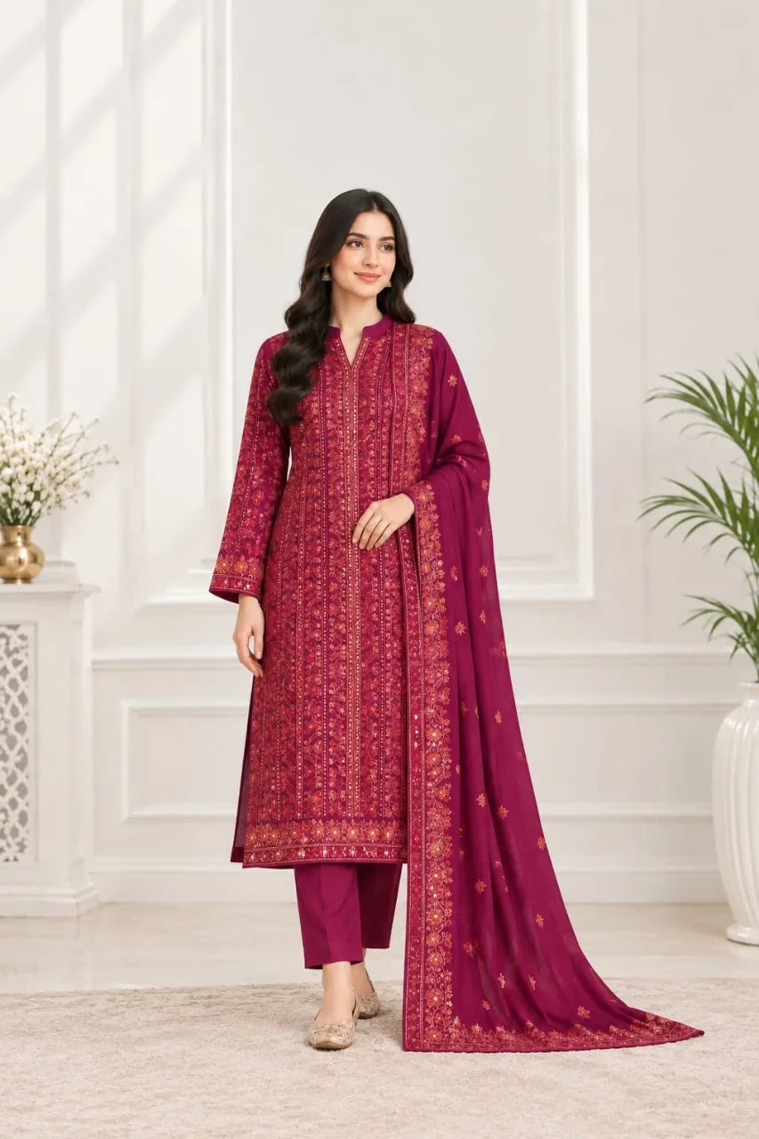 A woman models a traditional salwar kameez suit in deep magenta with intricate gold embroidery, paired with matching trousers and a long dupatta, set against a bright, modern interior background.