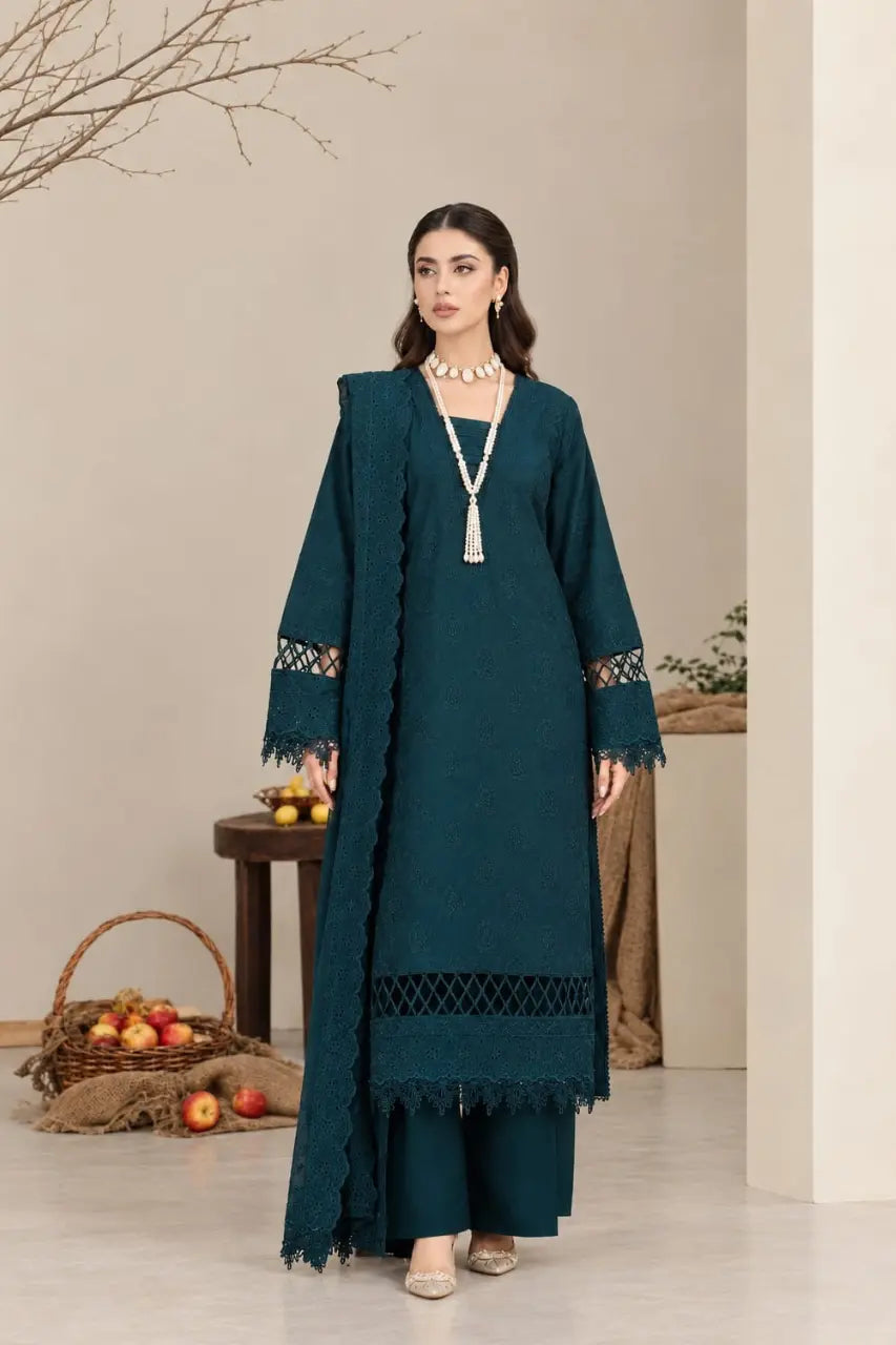 A woman modeling a dark teal three-piece traditional outfit with a long kurta, matching trousers, and a draped dupatta, styled with a tassel necklace in a warm neutral indoor setting.