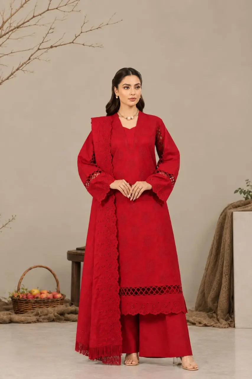 A woman stands indoors wearing a long red traditional outfit with a matching draped scarf, wide sleeves, and scalloped lace trim. The setting has a neutral beige wall, decorative branches, a basket of fruit, and draped fabric in the background.